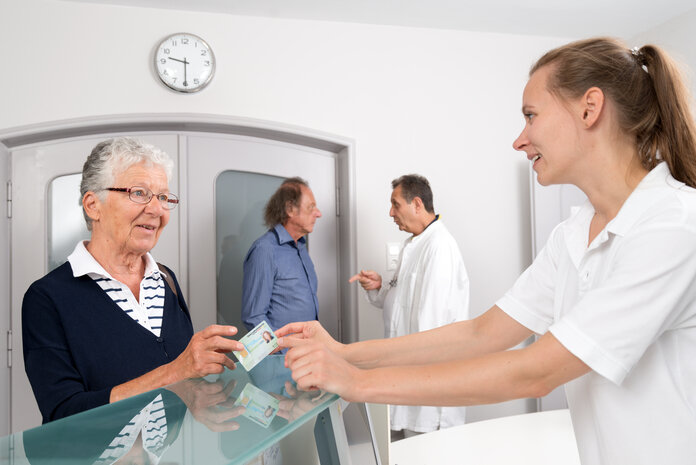 Arztpraxis: Empfang, © Krebsinformationsdienst, DKFZ; Foto: Tobias Schwerdt Patientin reicht einer Arzthelferin ihre Versichertenkarte am Empfang einer Arztpraxis. Im Hintergrund dpricht ein Arzt mit einem Patienten.